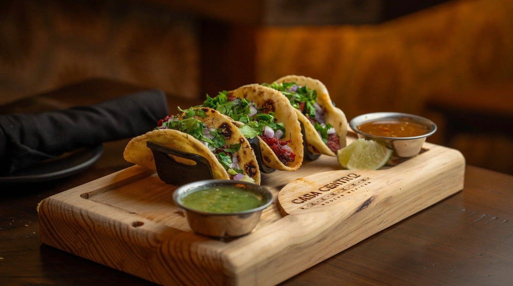 Three street-style tacos on a Casa Centro wooden serving board with red and green salsas and a lime wedge